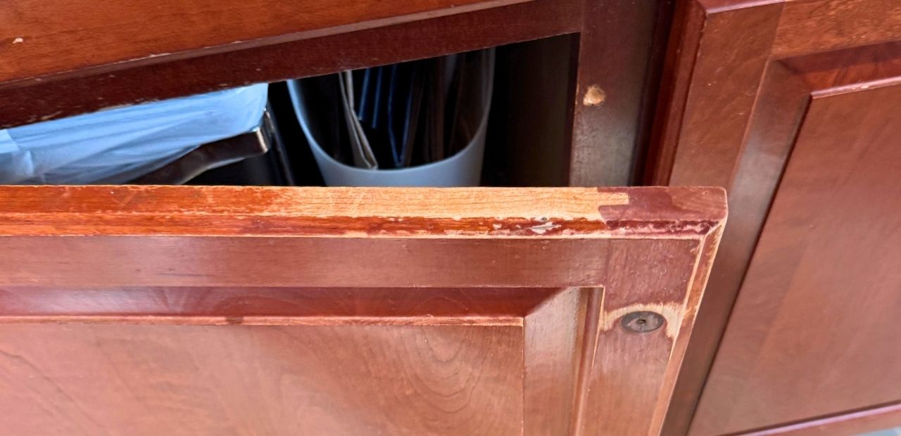 Before: Close-up of cherry kitchen door in Olney showing deep scratches, worn raw wood along the top edge, and missing finish.