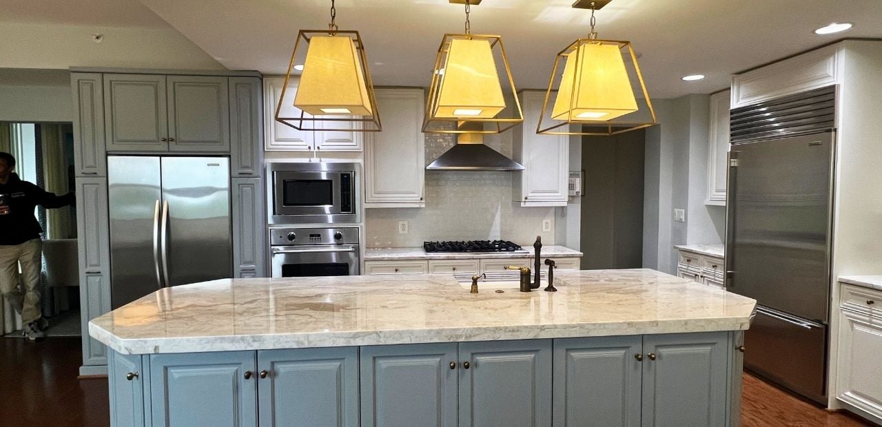 Before: Spacious North Bethesda kitchen with muted gray raised-panel cabinets, matching paneled refrigerator surrounds, blue-gray island, brushed-nickel hardware, and light stone countertop under three gold lantern pendants.