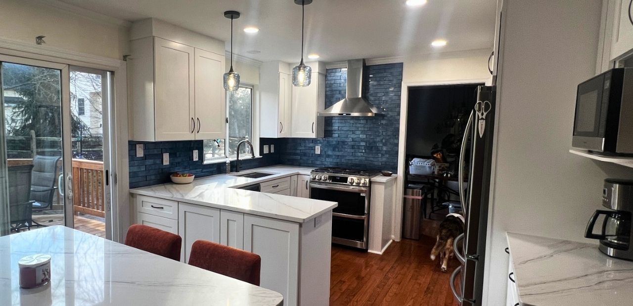 After: Two-tone white and gray Shaker kitchen in Olney with Calacatta quartz, blue glass backsplash, movable island, and stainless chimney hood delivering more storage.