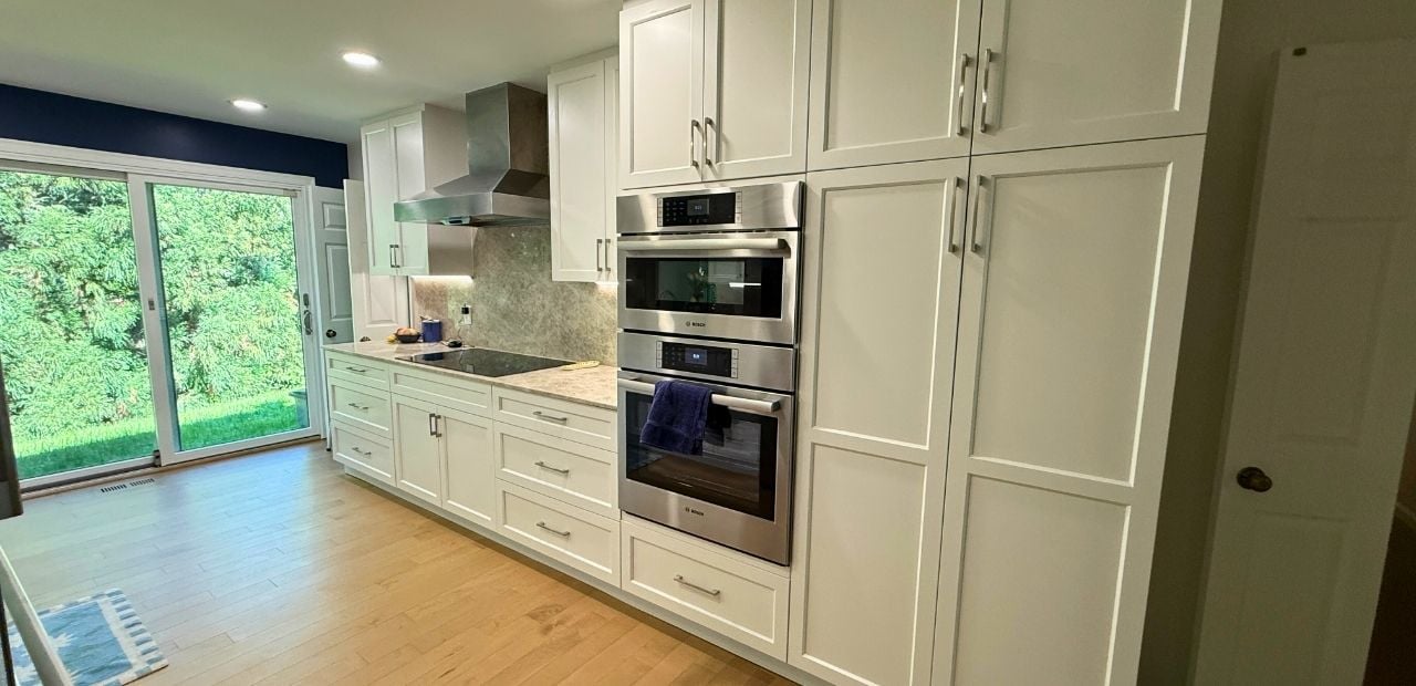 After: Bright white Shaker-style galley kitchen in North Bethesda with ceiling-height pantry towers, Bosch double wall ovens, stainless chimney range hood, quartzite countertop also installed as backsplash, warm hardwood flooring, and dimmable under-cabinet lighting.
