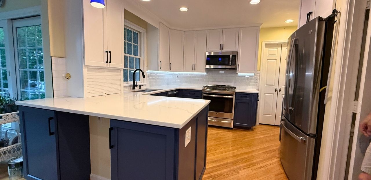 After: Rockville kitchen with beveled-Shaker doors—Satin White uppers, Van Deusen Blue bases and peninsula, one-level white quartz counters, matte-black hardware and faucet, subway-tile backsplash, and LED under-cab lighting.