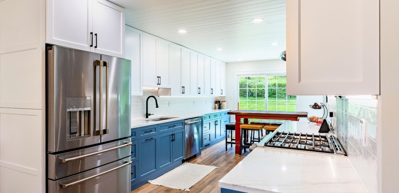 After: Bright galley kitchen featuring white Shaker uppers, navy Shaker lowers, matte-black hardware, white quartz counters, linear white backsplash, stainless appliances, shiplap ceiling with recessed lights, and a wood table island with stools.