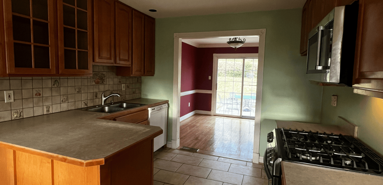 Before: Narrow galley kitchen with medium-tone oak cabinets, decorative fruit-tile backsplash, sage-green walls, laminate counters, white dishwasher, and view into a burgundy dining room.